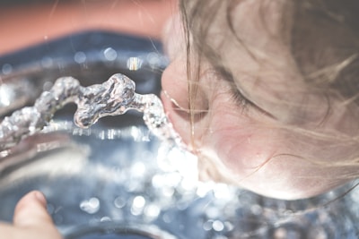 A close-up view of a young child drinking water from a fountain. The focus is on the child's face and the stream of water, with blurred background. The child's hair is slightly wet, indicating playfulness and refreshment.