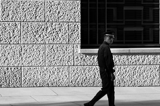 A person in uniform, possibly a security guard or police officer, walks alongside a textured wall. The wall has visible large blocks with a coarse surface. Shadows and light create strong contrasts, enhancing the texture of the surface.