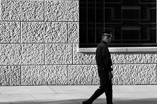 A person in uniform, possibly a security guard or police officer, walks alongside a textured wall. The wall has visible large blocks with a coarse surface. Shadows and light create strong contrasts, enhancing the texture of the surface.