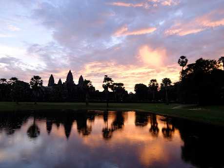 A silhouette of the iconic Angkor Wat temple complex is set against a vibrant sunrise or sunset sky, with hues of orange, pink, and purple. The reflective surface of a calm body of water in the foreground mirrors the sky and the temple's shadow, adding to the picturesque scenery. Palm trees are scattered around the temple, enhancing the tranquil atmosphere.