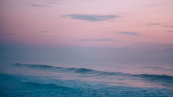 A serene ocean scene with gentle waves rolling towards the shore. The sky is painted in soft hues of pink and orange, suggesting either dawn or dusk. The overall appearance is calm and tranquil, with the horizon barely visible through the mist.