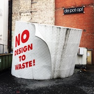 A large, sculptural installation resembling a roll of toilet paper. It is textured with small dot patterns and has bold red text stating 'NO DESIGN TO WASTE!' painted on it. The installation is positioned outdoors near a brick wall and a sign with the text 'de pot op!'. The ground is paved, and there is a metal container on the side.