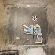 A piece of street art on a textured wall depicts a hand holding a flower. The hand is crafted from cutouts of newspaper print, adding a unique collage effect. The flower has simple, graphic styling with white petals and a red center, contrasting against the muted tones of the wall.
