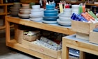 A wooden table displays multiple stacks of ceramic dishes in various colors, including white, blue, and gray. Beside the dishes, there are colorful rolled and stacked cloths in wooden crates. Below the table, woven baskets and more folded textiles are stored. The setup suggests a home goods or kitchenware store.
