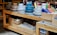 A wooden table displays multiple stacks of ceramic dishes in various colors, including white, blue, and gray. Beside the dishes, there are colorful rolled and stacked cloths in wooden crates. Below the table, woven baskets and more folded textiles are stored. The setup suggests a home goods or kitchenware store.