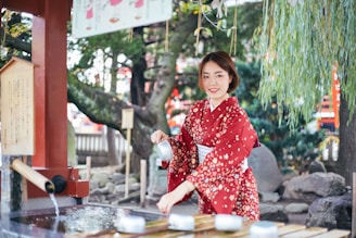 A woman wearing a red floral kimono is performing a traditional cleansing ritual at a water basin in a serene outdoor setting. She is holding a water ladle and smiling, surrounded by lush greenery and traditional Japanese architecture.
