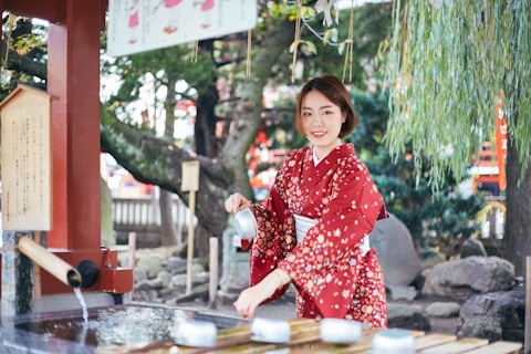 A woman wearing a red floral kimono is performing a traditional cleansing ritual at a water basin in a serene outdoor setting. She is holding a water ladle and smiling, surrounded by lush greenery and traditional Japanese architecture.