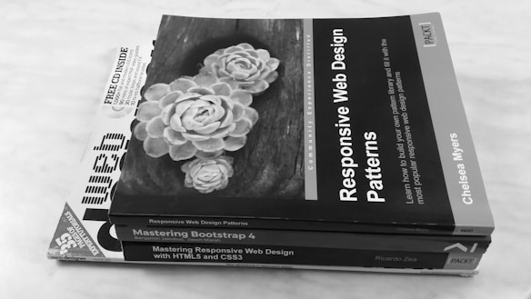 A stack of three web design books on a light surface. The top book features a cover with an image of white flowers and the title 'Responsive Web Design Patterns'. The second book is titled 'Mastering Bootstrap 4' and the third is 'Mastering Responsive Web Design with HTML5 and CSS3'.