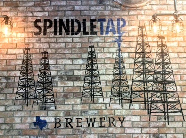 A brick wall features black stenciled oil derricks and industrial-style lighting fixtures. The name 'Spindletap' is prominently displayed in bold letters, with the word 'Brewery' below. A small blue silhouette of Texas is visible near the bottom.