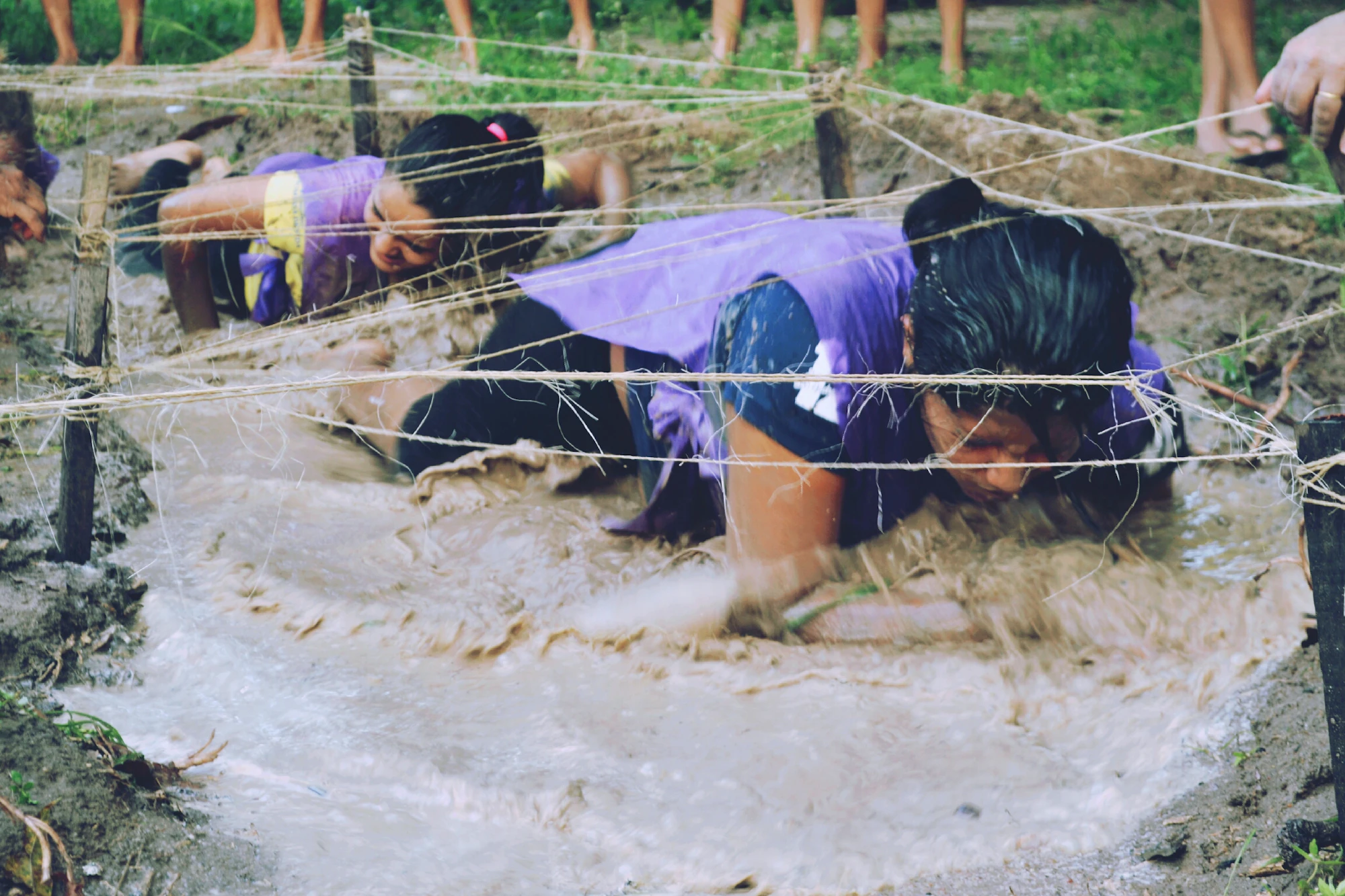 Obstacle-race competitors crawling under rope through mud