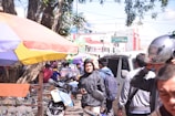 A busy street market with several people walking around. Vendors are set up under colorful umbrellas selling various items. A man dressed in a black jacket holds a smartphone, while another person adjusts their helmet. Scooters and motorcycles are parked nearby, and a bank building is visible in the background.