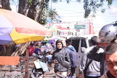 A busy street market with several people walking around. Vendors are set up under colorful umbrellas selling various items. A man dressed in a black jacket holds a smartphone, while another person adjusts their helmet. Scooters and motorcycles are parked nearby, and a bank building is visible in the background.