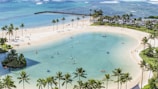 A tranquil beach scene featuring a serene lagoon surrounded by sandy shores. Several people are engaged in activities such as kayaking and paddleboarding on the calm water. Palm trees border the area, providing a tropical atmosphere. In the background, the ocean stretches to the horizon, with gentle waves visible. There is a small island with lush greenery in the lagoon, and a line of parked cars signifies a busy area nearby.