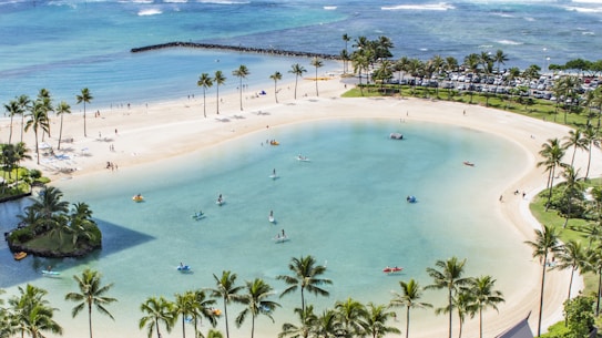 A tranquil beach scene featuring a serene lagoon surrounded by sandy shores. Several people are engaged in activities such as kayaking and paddleboarding on the calm water. Palm trees border the area, providing a tropical atmosphere. In the background, the ocean stretches to the horizon, with gentle waves visible. There is a small island with lush greenery in the lagoon, and a line of parked cars signifies a busy area nearby.