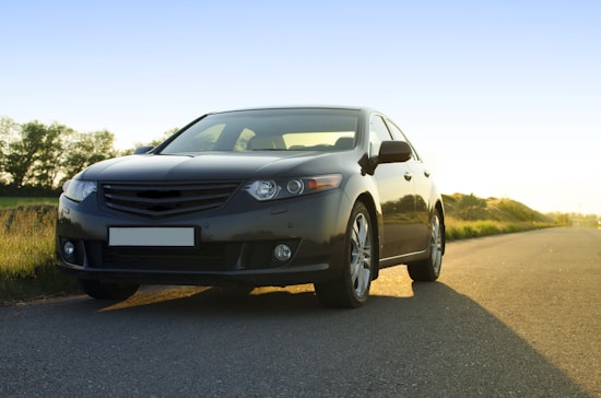 A sleek black car parked on a quiet road during sunset, with a clear sky and some greenery in the background. The light casts a warm glow, enhancing the car's polished surface.