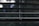Horizontal window blinds with thin cords hanging down in the center. Behind the blinds, a blurry, green-lit background can be seen, suggesting it's daylight outside.