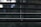 Horizontal window blinds with thin cords hanging down in the center. Behind the blinds, a blurry, green-lit background can be seen, suggesting it's daylight outside.