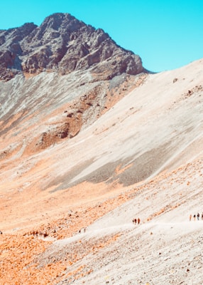 A rugged mountain landscape with a prominent rocky peak in the background and a gentle slope in the foreground. The terrain is mostly rocky with patches of beige and gray tones. A small group of people is walking along a winding path, adding a sense of scale and adventure.