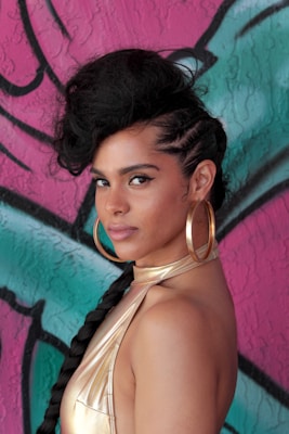 A person with an elaborate hairstyle featuring braids and a large, voluminous section on top. They are wearing gold hoop earrings and a metallic gold top, standing against a colorful graffiti wall with shades of pink and teal.