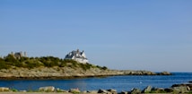 A coastal scene with a large house situated on a rocky promontory. The house is surrounded by greenery and overlooks a calm sea. The sky is clear, enhancing the tranquil atmosphere.