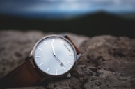A wristwatch with a minimalist design features a white face with slender metallic markings for the hours and a brown leather strap. It is placed on a rough, textured stone surface, with a blurred background suggesting an outdoor setting.