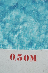 Clear blue water of a swimming pool is visible with a tiled floor. The edge of the pool shows a measurement marking of 0.50M in red stencil on the white surface.