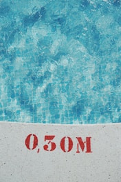 Clear blue water of a swimming pool is visible with a tiled floor. The edge of the pool shows a measurement marking of 0.50M in red stencil on the white surface.