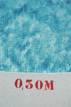 Clear blue water of a swimming pool is visible with a tiled floor. The edge of the pool shows a measurement marking of 0.50M in red stencil on the white surface.