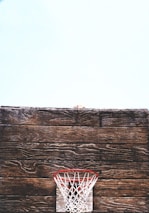 A rustic wooden basketball backboard is prominently displayed with an old-fashioned red and white net situated against a light blue sky. The worn texture of the wood adds a vintage feel to the scene.