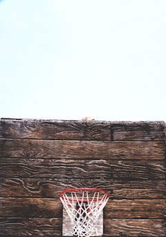 A rustic wooden basketball backboard is prominently displayed with an old-fashioned red and white net situated against a light blue sky. The worn texture of the wood adds a vintage feel to the scene.