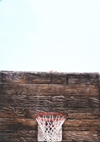 A rustic wooden basketball backboard is prominently displayed with an old-fashioned red and white net situated against a light blue sky. The worn texture of the wood adds a vintage feel to the scene.