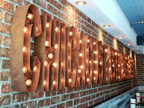 A long brick wall featuring a large, metallic sign with cut-out letters that have light bulbs inside, illuminating the area. The sign is reminiscent of a classic, rustic style. The ceiling features pot lights, providing additional illumination.