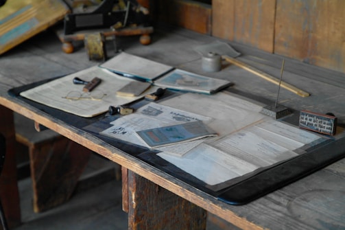 An old wooden desk is filled with various vintage items including papers, an ink well with a pen, a small calendar, a pair of eyeglasses, and a wooden stamp. The items are spread out in an organized yet cluttered manner, evoking a sense of historical or archival work.