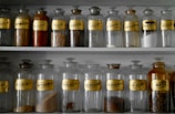 A collection of vintage glass apothecary jars is neatly arranged on shelves. Each jar has a gold label with black lettering, indicating different substances. Some jars contain powders or herbs, while others appear empty.