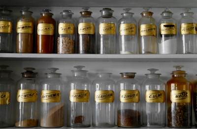 A collection of vintage glass apothecary jars is neatly arranged on shelves. Each jar has a gold label with black lettering, indicating different substances. Some jars contain powders or herbs, while others appear empty.