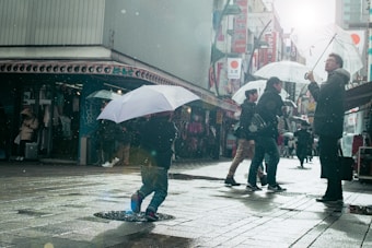 A rainy street scene in an urban area features several people walking with umbrellas. The damp ground reflects the city lights and signage in the background. Some pedestrians appear to be in mid-conversation or looking at something as they move along the sidewalk.
