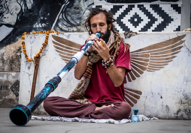A person with long hair and a beard is seated cross-legged, playing a didgeridoo. They wear a red shirt and have several bracelets on their wrist. The wall behind them is decorated with wing-like artwork and patterns in black and white. A garland hangs nearby.