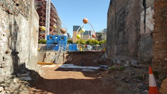 A construction site with exposed brick walls on either side and a cleared dirt area in the center. In the background, there are several colorful, spherical sculptures atop thin poles, with blue shipping containers and fencing surrounding the area. A tall building with metal scaffolding is partially visible behind the sculptures. Orange construction cones are placed on the ground, and various construction materials are scattered around.