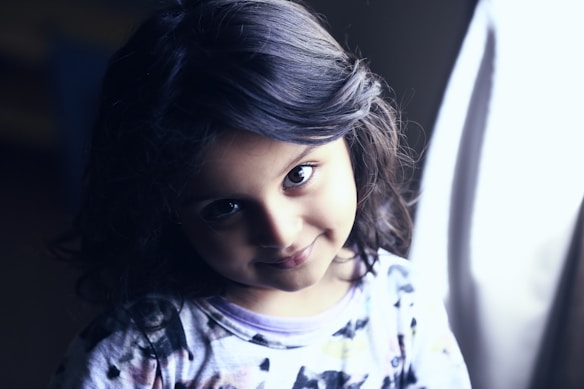 A young girl with dark hair is looking at the camera with a gentle smile. Her hair is styled in soft waves, and she wears a patterned shirt. Soft lighting illuminates her face, creating a focused and intimate atmosphere.