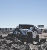 A rugged off-road vehicle is positioned on rocky terrain against a clear blue sky. The vehicle appears sturdy and equipped for challenging environments, with visible modifications like large tires and additional lights. A fence and construction site can be seen in the background, suggesting an industrial setting.