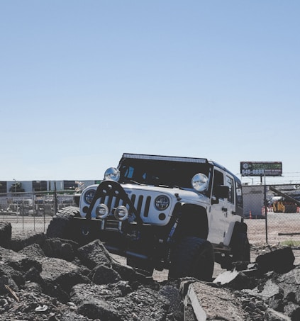 A rugged off-road vehicle is positioned on rocky terrain against a clear blue sky. The vehicle appears sturdy and equipped for challenging environments, with visible modifications like large tires and additional lights. A fence and construction site can be seen in the background, suggesting an industrial setting.