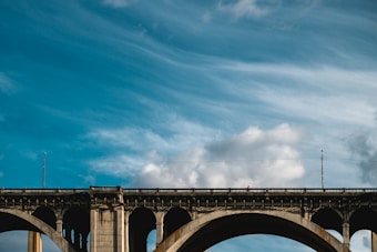 An arch bridge stretches across the image with a bright blue sky and wispy clouds above. The bridge is made of concrete with strong pillars and railings visible along the top.
