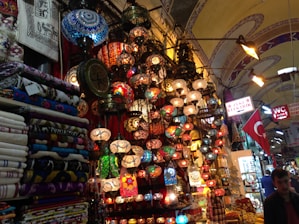A vibrant marketplace scene with an array of colorful mosaic lamps hanging from the ceiling, each with intricate patterns. Nearby, stacks of patterned textiles and fabrics are neatly folded. The environment is bustling with signs in Turkish language and a Turkish flag displayed prominently. The setting suggests a rich cultural ambiance enriched by traditional artisan craftsmanship.