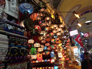 A vibrant marketplace scene with an array of colorful mosaic lamps hanging from the ceiling, each with intricate patterns. Nearby, stacks of patterned textiles and fabrics are neatly folded. The environment is bustling with signs in Turkish language and a Turkish flag displayed prominently. The setting suggests a rich cultural ambiance enriched by traditional artisan craftsmanship.