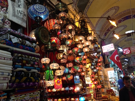 A vibrant marketplace scene with an array of colorful mosaic lamps hanging from the ceiling, each with intricate patterns. Nearby, stacks of patterned textiles and fabrics are neatly folded. The environment is bustling with signs in Turkish language and a Turkish flag displayed prominently. The setting suggests a rich cultural ambiance enriched by traditional artisan craftsmanship.