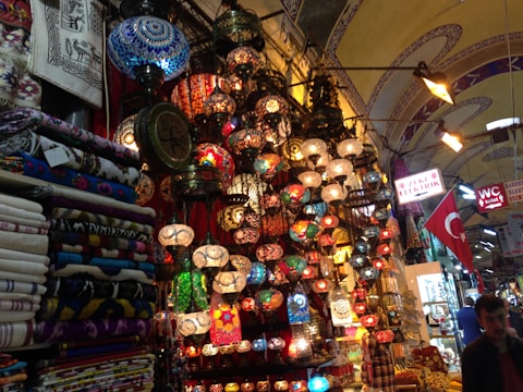A vibrant marketplace scene with an array of colorful mosaic lamps hanging from the ceiling, each with intricate patterns. Nearby, stacks of patterned textiles and fabrics are neatly folded. The environment is bustling with signs in Turkish language and a Turkish flag displayed prominently. The setting suggests a rich cultural ambiance enriched by traditional artisan craftsmanship.