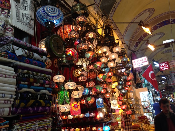 A vibrant marketplace scene with an array of colorful mosaic lamps hanging from the ceiling, each with intricate patterns. Nearby, stacks of patterned textiles and fabrics are neatly folded. The environment is bustling with signs in Turkish language and a Turkish flag displayed prominently. The setting suggests a rich cultural ambiance enriched by traditional artisan craftsmanship.