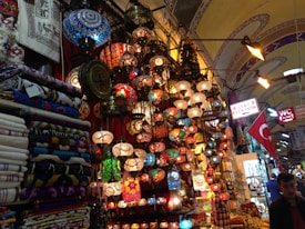 A vibrant marketplace scene with an array of colorful mosaic lamps hanging from the ceiling, each with intricate patterns. Nearby, stacks of patterned textiles and fabrics are neatly folded. The environment is bustling with signs in Turkish language and a Turkish flag displayed prominently. The setting suggests a rich cultural ambiance enriched by traditional artisan craftsmanship.