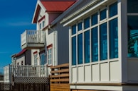 A row of modern homes with large windows and small balconies. The buildings showcase a clean architectural design with white and red detailing. The sky is clear and blue, illuminating the facade of the houses.