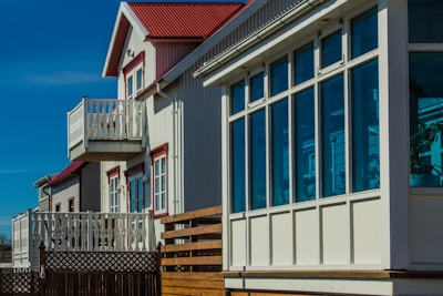 A row of modern homes with large windows and small balconies. The buildings showcase a clean architectural design with white and red detailing. The sky is clear and blue, illuminating the facade of the houses.